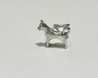 May include: A silver-colored charm in the shape of a dog, likely a Shiba Inu, with detailed fur texture. The charm has a loop at the top for attachment to a necklace or bracelet. The dog is standing, facing left.