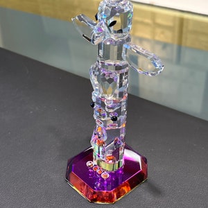May include: A crystal totem pole figurine with a purple and orange base. The totem pole is made of clear crystal and features a variety of animal and human figures.