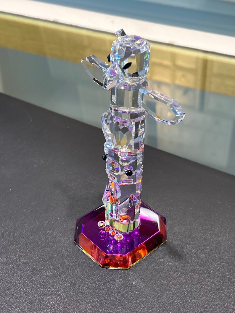 May include: A crystal totem pole figurine with a purple and orange base. The totem pole is made of clear crystal and features a variety of animal and human figures.