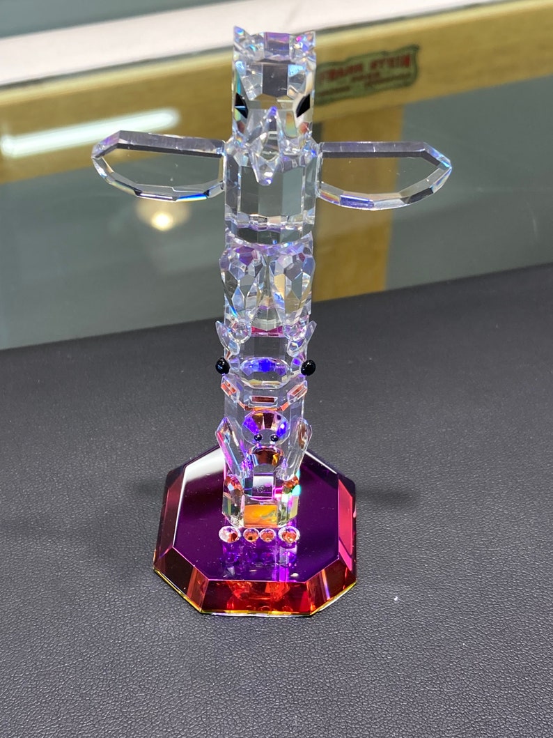 May include: A crystal totem pole figurine with a clear glass base and a purple and red iridescent finish. The totem pole features a bird with outstretched wings and a bear with black eyes.