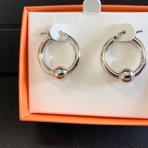 Op de afbeelding: Een paar zilveren creolen met een balbedel. De oorbellen zitten in een witte doos met een oranje deksel.