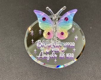 Könnte beinhalten: Eine transparente Glas-Schmetterlingsfigur mit schillernden Flügeln in Lila, Blau und Gelb, verziert mit lila Edelsteinen. Die Figur steht auf einem runden, verspiegelten Sockel mit dem Text "Butterflies appear when Angels are near."