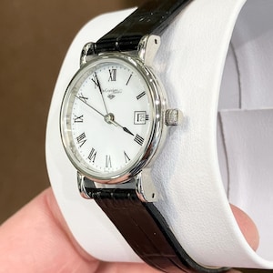 May include: A silver-toned wristwatch with a white face, black Roman numerals, and a date display at the 3 o'clock position. The watch has a black leather band and a small crown. The watch is displayed on a white surface.