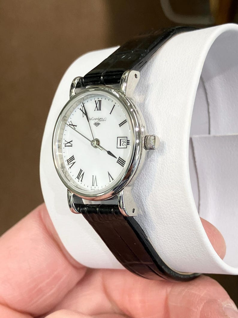 May include: A silver-toned wristwatch with a white face, black Roman numerals, and a date display at the 3 o'clock position. The watch has a black leather band and a small crown. The watch is displayed on a white surface.