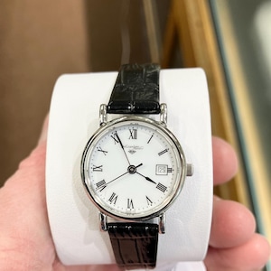 May include: A silver-toned wristwatch with a white face, black Roman numeral hour markers, and a date display at the 3 o'clock position. The watch has a black leather band and is held on a white display. The watch face reads "Grandeur".