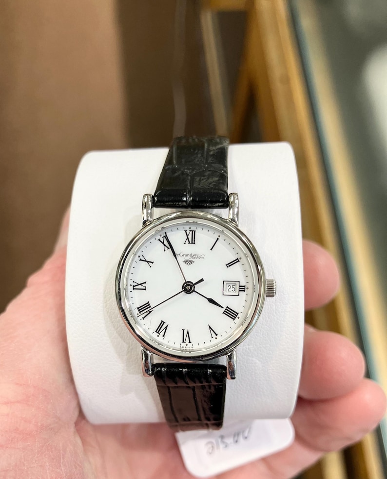 May include: A silver-toned wristwatch with a white face, black Roman numeral hour markers, and a date display at the 3 o'clock position. The watch has a black leather band and is held on a white display. The watch face reads "Grandeur".