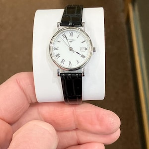 May include: A silver-toned wristwatch with a white face, black Roman numerals, and a date window at the 3 o'clock position. The watch has a black leather band. The watch is displayed on a white stand.