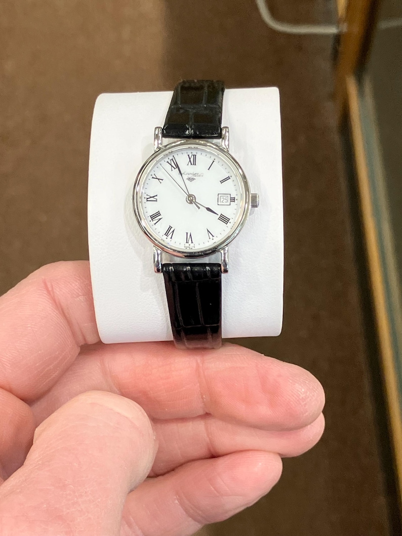 May include: A silver-toned wristwatch with a white face, black Roman numerals, and a date window at the 3 o'clock position. The watch has a black leather band. The watch is displayed on a white stand.