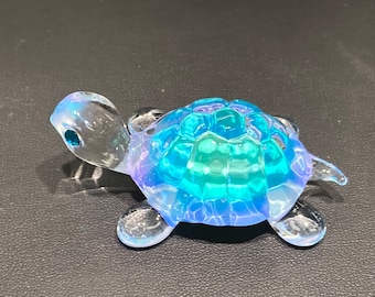 May include: A small, iridescent glass turtle figurine. The turtle's shell is a mosaic of blues and greens, while its head and limbs are clear. The figurine has a small, blue eye and is resting on a dark surface.