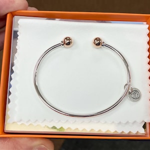 Könnte beinhalten: Ein silbernes Armreif-Armband mit zwei roségoldenen Akzentkugeln und einem kleinen silbernen Anhänger. Das Armband wird in einer weißen Box mit orangefarbenem Rand präsentiert.