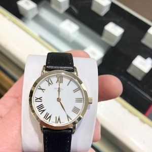 May include: A gold-tone watch with a white face and black leather strap. The watch face has Roman numeral hour markers and a gold-tone second hand.
