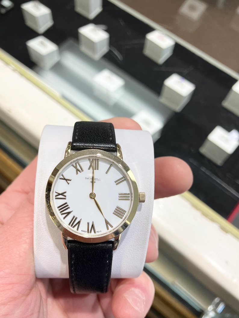 May include: A gold-tone watch with a white face and black leather strap. The watch face has Roman numeral hour markers and a gold-tone second hand.