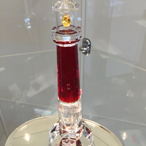 May include: A red crystal lighthouse figurine with a clear glass top and a yellow crystal light. The base of the lighthouse is clear glass with a red crystal center.