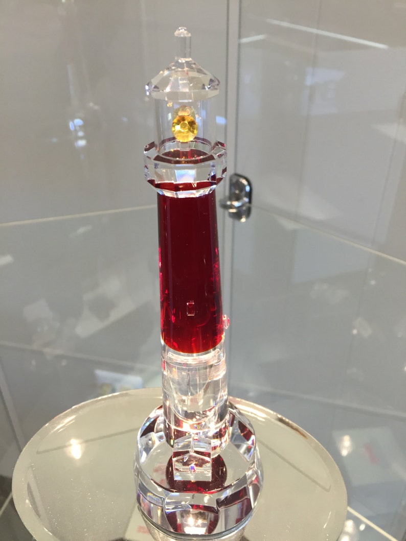 May include: A red crystal lighthouse figurine with a clear glass top and a yellow crystal light. The base of the lighthouse is clear glass with a red crystal center.