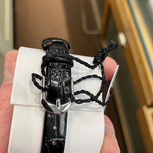 May include: A black leather watch band with a silver buckle is wrapped around a white cushion. The band is secured with a black cord and a tag. The watch band has a textured, crocodile-like pattern.