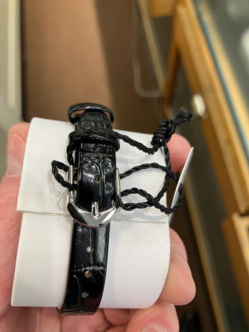 May include: A black leather watch band with a silver buckle is wrapped around a white cushion. The band is secured with a black cord and a tag. The watch band has a textured, crocodile-like pattern.