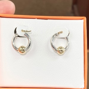 Puede incluir: Un par de pendientes de aro de plata con un acento de bola dorada. Los pendientes se muestran en una caja blanca con un borde naranja.
