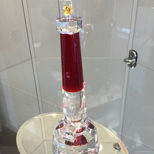May include: A red and clear glass lighthouse figurine with a small yellow crystal at the top. The figurine is on a clear glass base.