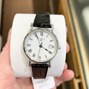May include: A silver-toned wristwatch with a white face, black Roman numerals, and a date display at the 3 o'clock position. The watch has a black leather band and is held on a white display. The watch face has the brand name "Casandra" printed on it.