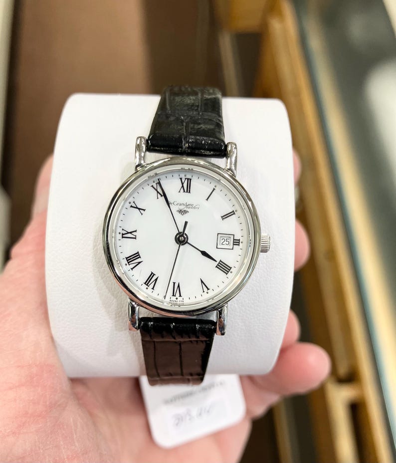 May include: A silver-toned wristwatch with a white face, black Roman numerals, and a date display at the 3 o'clock position. The watch has a black leather band and is held on a white display. The watch face has the brand name "Casandra" printed on it.