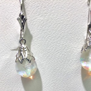 May include: A pair of silver-toned drop earrings. Each earring features a decorative top with a teardrop-shaped, iridescent, clear stone. The stones reflect a rainbow of colors. The earrings are displayed against a white background.