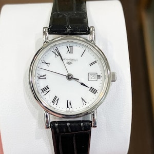 May include: A silver-toned wristwatch with a white face and black Roman numerals. The watch has a date display at the 3 o'clock position and a black leather band. The watch face reads "Grandeur Jewelers".