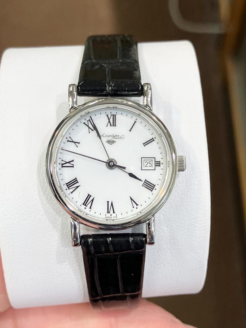 May include: A silver-toned wristwatch with a white face and black Roman numerals. The watch has a date display at the 3 o'clock position and a black leather band. The watch face reads "Grandeur Jewelers".