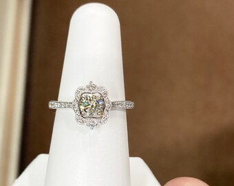 May include: A silver engagement ring with a round, clear center stone surrounded by smaller diamonds. The ring is set in a delicate, floral-inspired design.