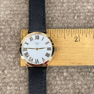 May include: A gold-tone wristwatch with a white face and black Roman numeral hour markers. The watch has a black leather strap and is displayed on a wooden ruler.