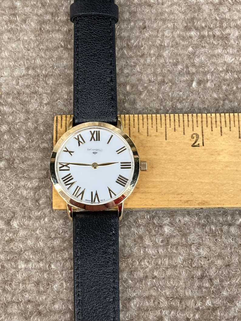 May include: A gold-tone wristwatch with a white face and black Roman numeral hour markers. The watch has a black leather strap and is displayed on a wooden ruler.