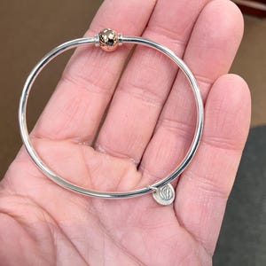 Op de afbeelding: Een zilveren armband met een ros&eacute;gouden balbedel en een zilveren bedel met een logo.