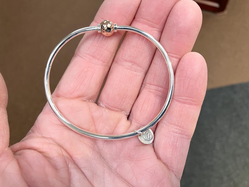 Op de afbeelding: Een zilveren armband met een ros&eacute;gouden balbedel en een zilveren bedel met een logo.