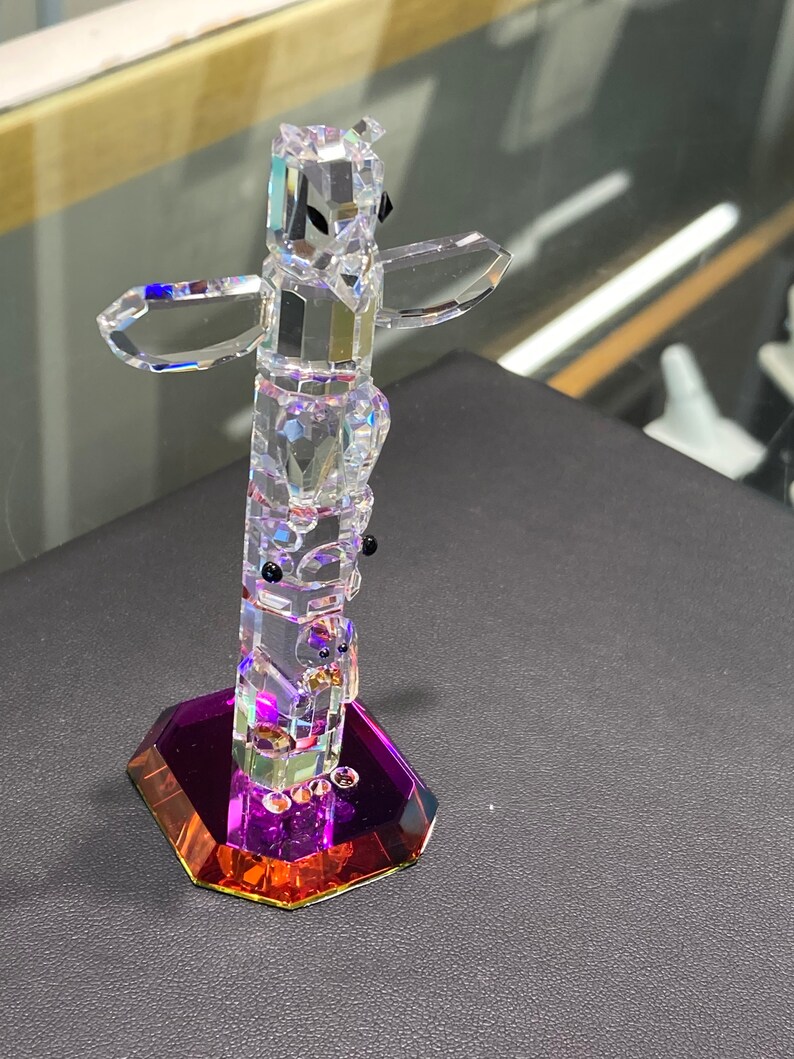 May include: A crystal totem pole figurine with a purple and orange base. The totem pole is made of clear crystal and features a variety of animal figures.