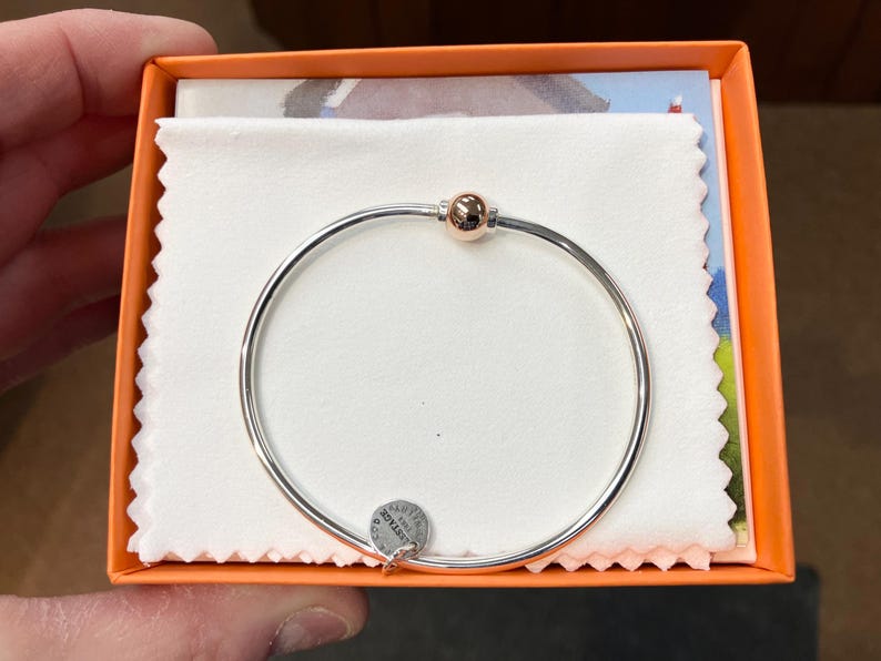 Op de afbeelding: Een zilveren armband met een ros&eacute;gouden kraalbedel. De armband zit in een kleine oranje doos met een witte stoffen voering. De doos heeft een wit label met het woord "STERLING" erop.