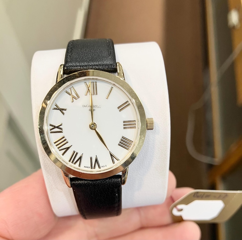 May include: A gold-tone wristwatch with a white face and black leather strap. The watch has Roman numeral hour markers and a gold-tone second hand.