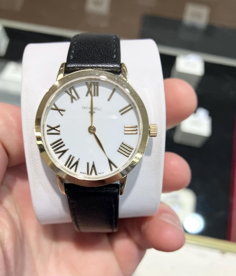 May include: A gold-tone watch with a white face and black leather strap. The watch has Roman numeral hour markers and a gold-tone second hand.
