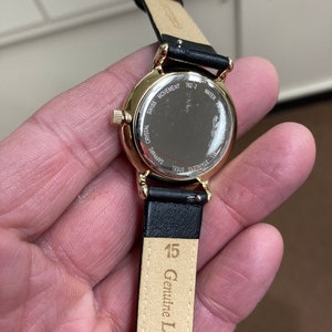 May include: A gold-tone wristwatch with a black leather strap. The watch face is protected by a sapphire crystal and the back of the watch case is engraved with "Swiss Movement", "Sapphire Crystal", "Stainless Steel", "762-2", and "Water Resistant". The strap is labelled "Genuine Leather" and "15".