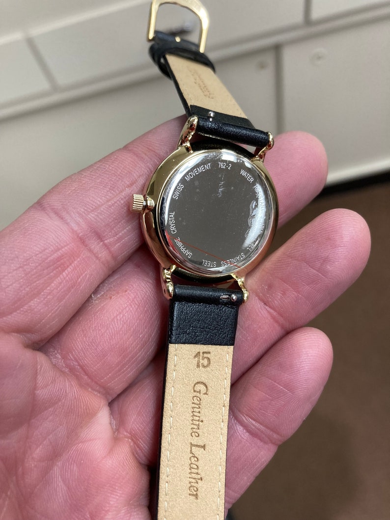 May include: A gold-tone wristwatch with a black leather strap. The watch face is protected by a sapphire crystal and the back of the watch case is engraved with "Swiss Movement", "Sapphire Crystal", "Stainless Steel", "762-2", and "Water Resistant". The strap is labelled "Genuine Leather" and "15".