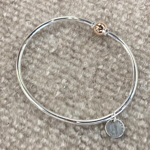 Op de afbeelding: Een zilveren armband met een gouden bolvormige bedel en een zilveren schijfbedel. De schijfbedel heeft een inscriptie.