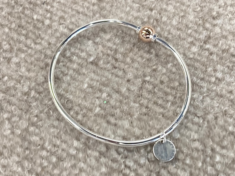 Op de afbeelding: Een zilveren armband met een gouden bolvormige bedel en een zilveren schijfbedel. De schijfbedel heeft een inscriptie.