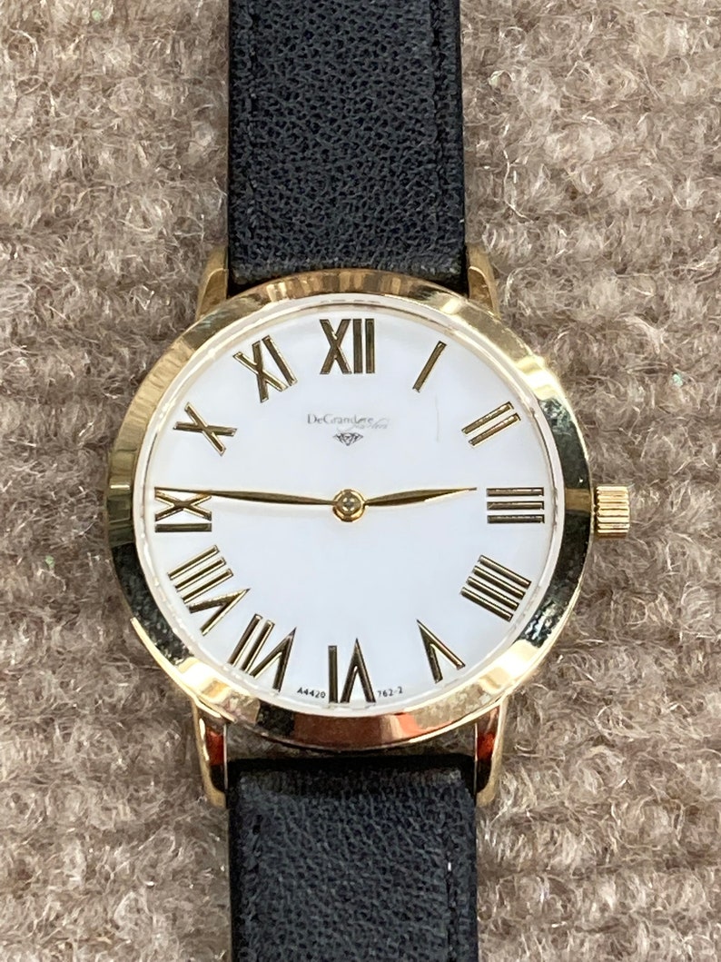 May include: A gold-tone wristwatch with a white face and black leather strap. The watch face has Roman numeral hour markers and a DeGramere logo.