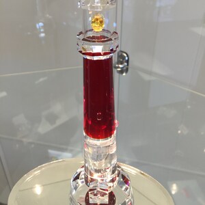 May include: A red crystal lighthouse figurine with a clear glass top and a yellow crystal light. The lighthouse is on a clear glass base.