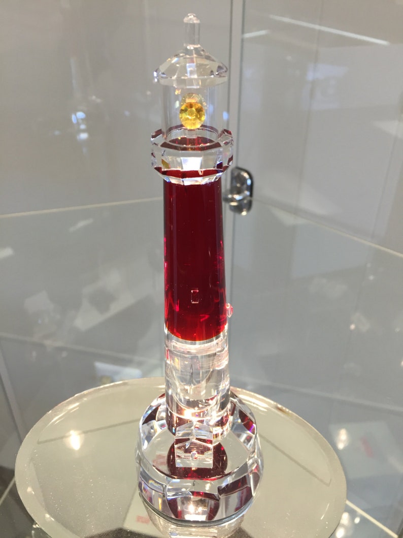 May include: A red crystal lighthouse figurine with a clear glass top and a yellow crystal light. The lighthouse is on a clear glass base.