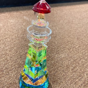 Pode incluir: Uma figura de farol de cristal com um telhado vermelho e um corpo de vidro transparente. O farol é decorado com um arco-íris de cores, incluindo verde, amarelo, azul e dourado.