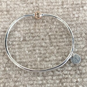 Op de afbeelding: Een zilveren armband met een gouden bolvormige bedel en een zilveren tag met het woord "Sterling" erop.