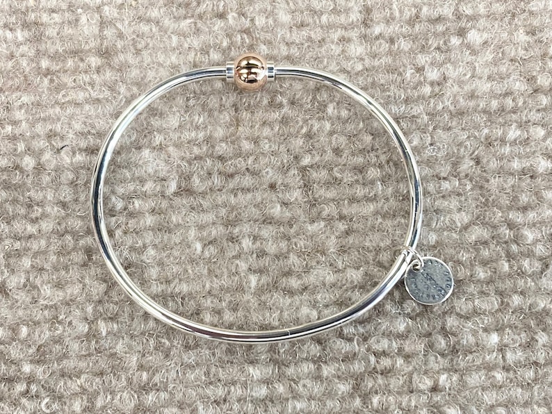Op de afbeelding: Een zilveren armband met een gouden bolvormige bedel en een zilveren tag met het woord "Sterling" erop.