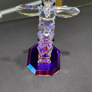 May include: A crystal totem pole figurine with a purple base. The totem pole is made of clear crystal and features a bird with outstretched wings at the top.