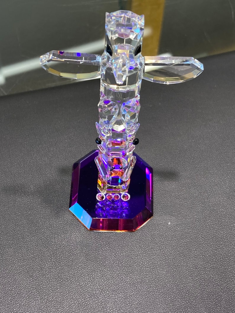 May include: A crystal totem pole figurine with a purple base. The totem pole is made of clear crystal and features a bird with outstretched wings at the top.