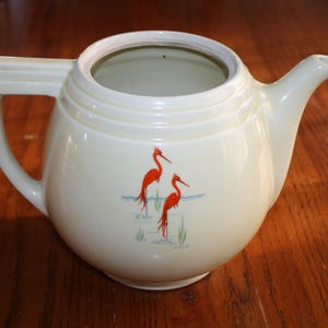 May include: A white ceramic teapot with a rounded body and a lid. The teapot has a handle and a spout. The teapot is decorated with two red birds with long legs and beaks.