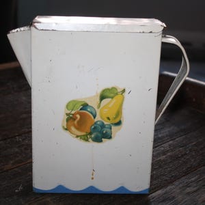 May include: White metal pitcher with a blue and white trim. The pitcher has a fruit design with a pear, peach, and grapes.